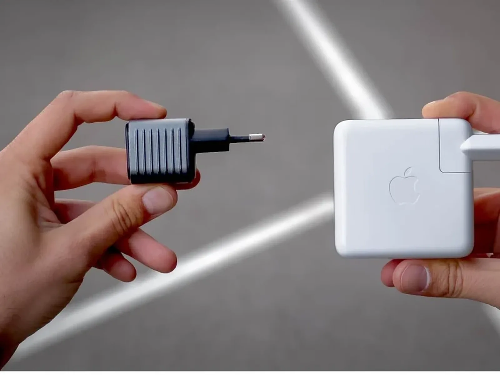 Two hands holding the Supertiny charger and an Apple MacBook charger side by side.